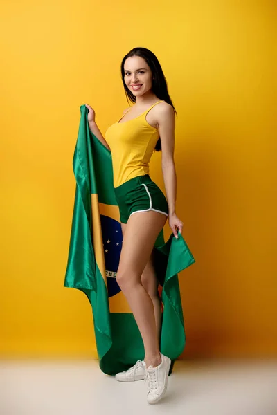 Allegro tifoso di calcio femminile in pantaloncini con bandiera brasiliana su giallo — Foto stock