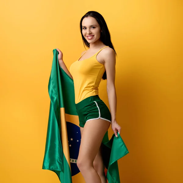 Attractive smiling football fan in shorts holding brazilian flag on yellow — Stock Photo