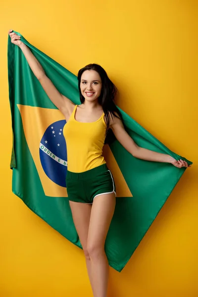 Cheerful female football fan in shorts holding brazilian flag on yellow — Stock Photo