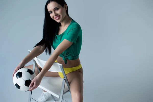 Cheerful sexy girl holding football ball while sitting on chair on grey — Stock Photo