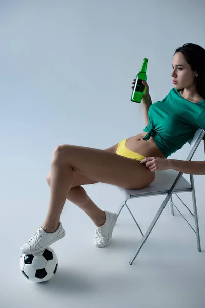 Attraktiver sexy Fußballfan mit Ball, der eine Flasche Bier hält, während er auf einem Stuhl auf grau sitzt — Stockfoto
