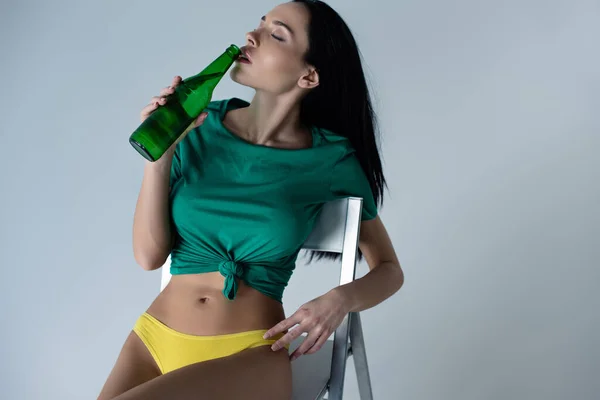 Sexy mulher beber cerveja enquanto sentado na cadeira isolado no cinza — Fotografia de Stock