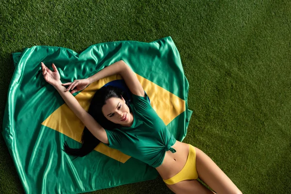 Vue du haut de fille sexy heureuse couché sur le drapeau brésilien sur l'herbe verte — Photo de stock