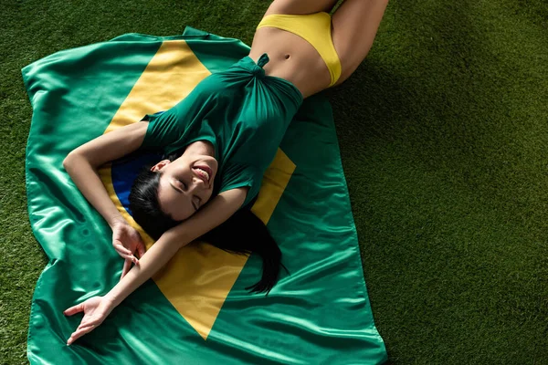 Top view of smiling sexy girl lying on brazilian flag on green grass — Stock Photo