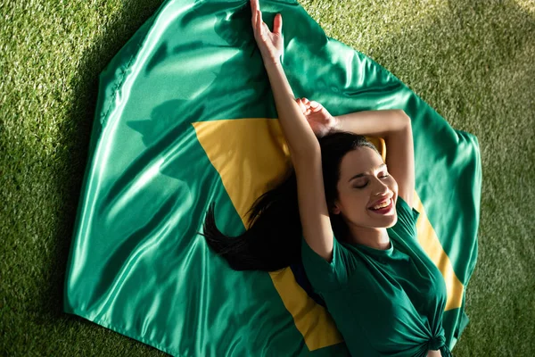 Lächelndes sexy Mädchen liegt auf brasilianischer Flagge auf grünem Gras — Stockfoto