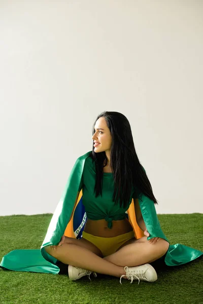 Sonriente ventilador de fútbol sexy sentado con bandera brasileña en hierba verde aislado en gris - foto de stock