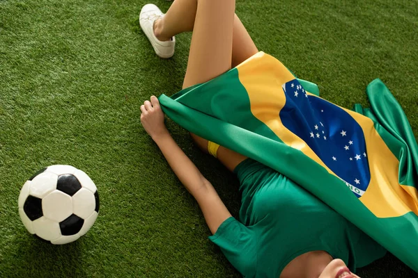 Vista cortada de menina sexy deitado com bandeira brasileira e bola de futebol na grama verde — Fotografia de Stock
