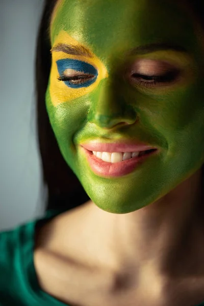 Attraktive, fröhliche Frau mit bemalter brasilianischer Flagge im Gesicht isoliert auf grau — Stockfoto
