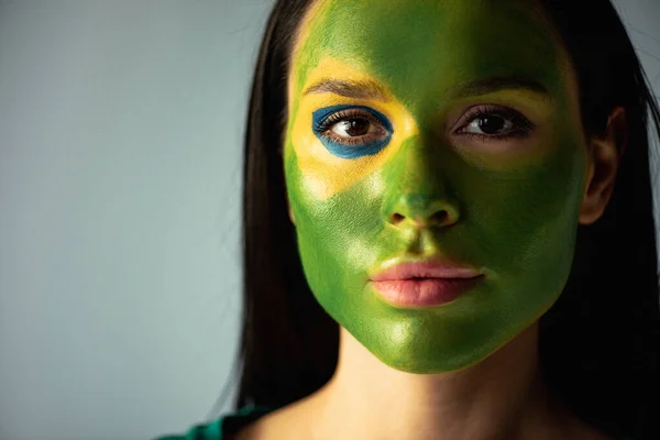 Beautiful female football fan with painted brazilian flag on face isolated on grey — Stock Photo