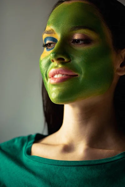 Attractive smiling woman with painted brazilian flag on face  isolated on grey — Stock Photo