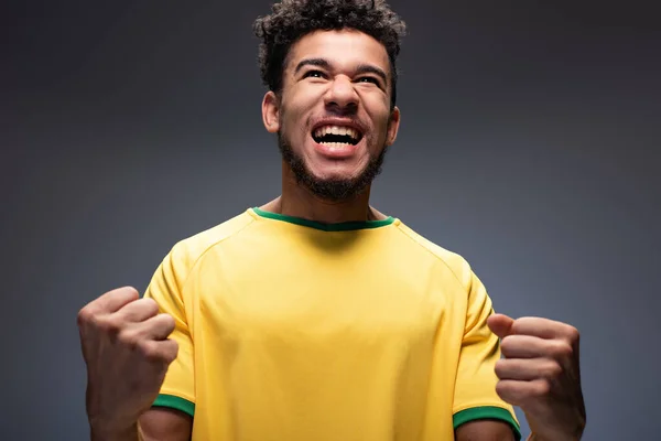 Возбужденный африканский американский футбольный фанат в желтой футболке, кричащий и жестикулирующий на сером — стоковое фото