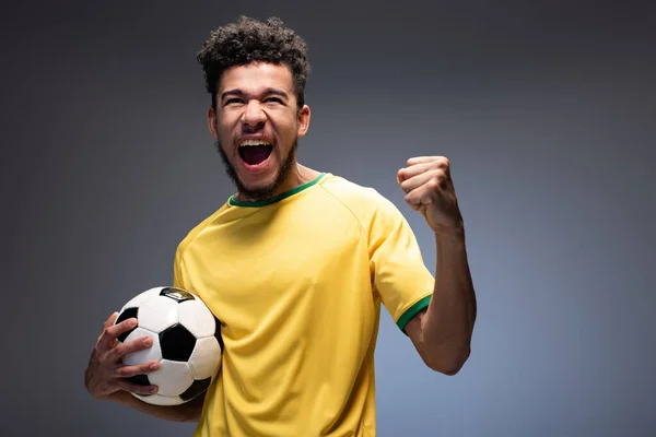 Fan de football afro-américain masculin excité en t-shirt jaune tenant le ballon sur gris — Photo de stock