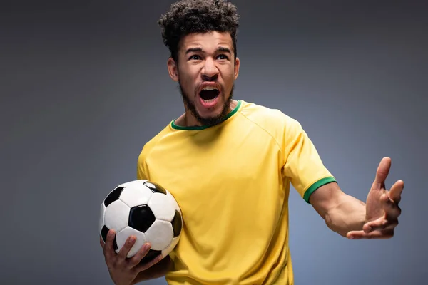Irritado fã de futebol americano africano em camiseta amarela gritando e segurando bola no cinza — Fotografia de Stock
