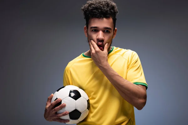 Frustrado fanático del fútbol afroamericano en camiseta amarilla sosteniendo la pelota en gris - foto de stock