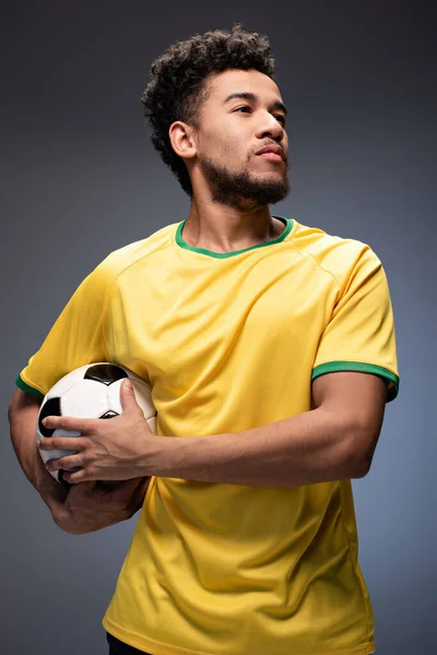 Fier fan de football afro-américain en t-shirt jaune tenant le ballon sur gris — Photo de stock