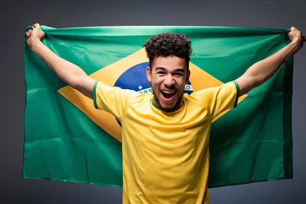 Aufgeregter afrikanisch-amerikanischer Fußballfan schreit und hält brasilianische Flagge auf grau — Stockfoto