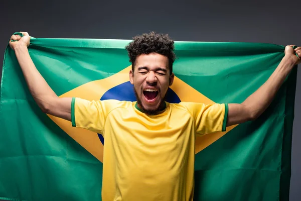 Emotional african american football fan shouting and holding brazilian flag on grey — Stock Photo