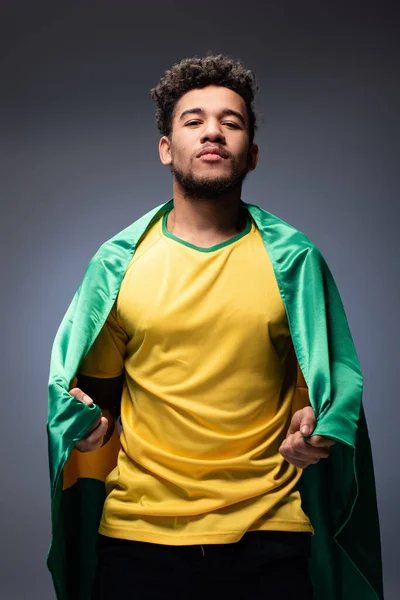 Fier fan de football afro-américain enveloppé dans le drapeau brésilien sur gris — Photo de stock