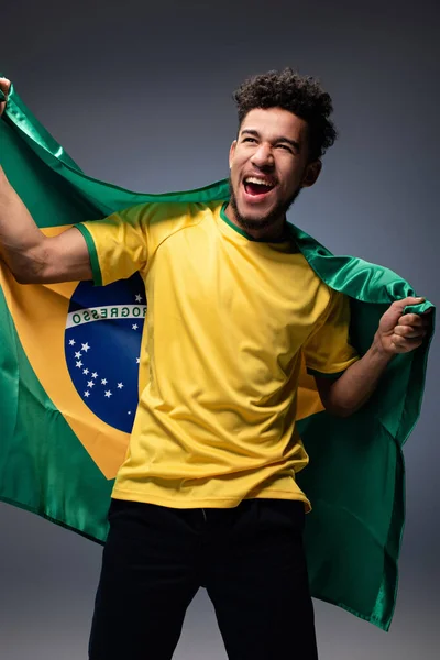 Схвильований афроамериканський футбольний фанат тримає бразильський прапор на сірому — стокове фото