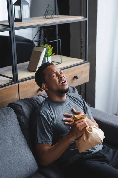 Homem afro-americano estressado segurando saco de papel enquanto tendo ataque de pânico em casa — Fotografia de Stock