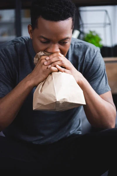 Lonely african american man breathing with paper bag while having panic attack at home — Stock Photo