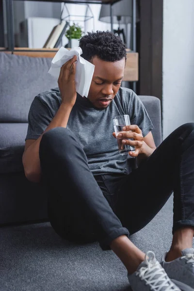 Stressed sweaty african american man holding glass of water and napkins at home — Stock Photo