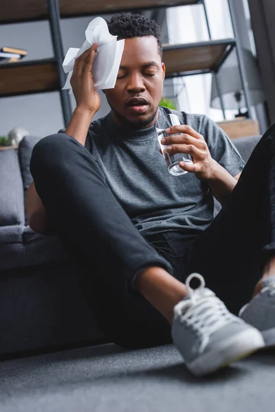 Sudado afroamericano hombre sosteniendo vaso de agua y servilletas después de ataque de pánico en casa - foto de stock