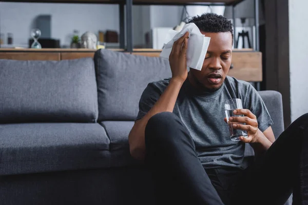 Sudado afroamericano hombre sosteniendo vaso de agua y servilletas después de ataque de pánico en casa - foto de stock