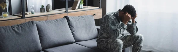 Panoramic shot of sad african american soldier in military uniform suffering from PTSD at home — Stock Photo