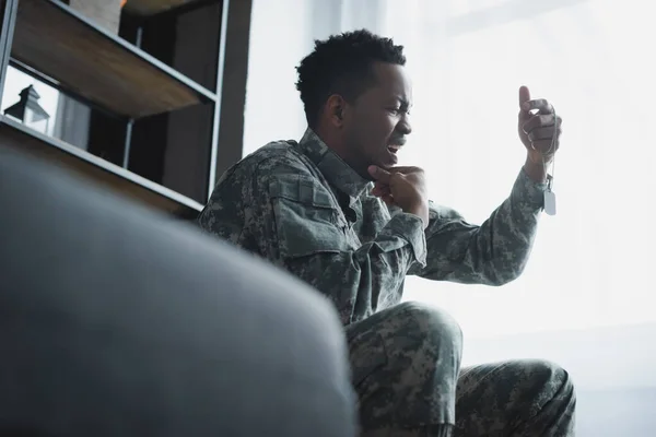 Estresado soldado afroamericano llorando y sosteniendo la insignia del ejército en casa, sufriendo de estrés postraumático - foto de stock
