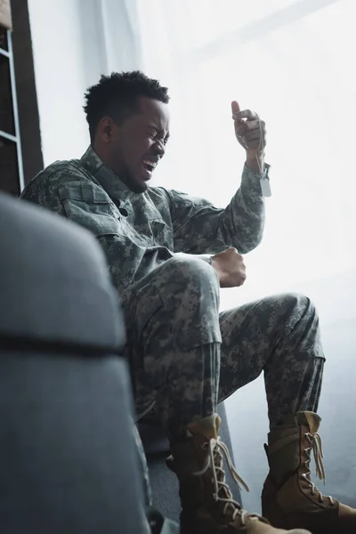 Soldado afroamericano estresado gritando y sosteniendo la insignia del ejército en casa, sufriendo de PTSD - foto de stock