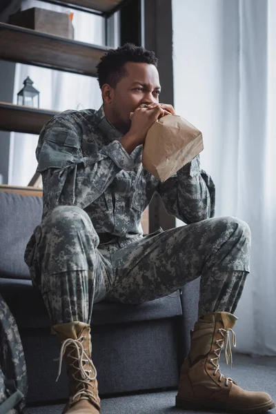 African american soldier in military uniform breathing with paper bag while having panic attack at home — Stock Photo