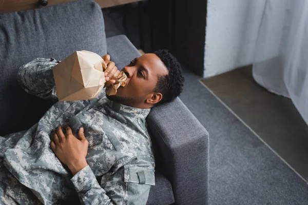 Stressed african american soldier in military uniform breathing with paper bag while having panic attack at home — Stock Photo