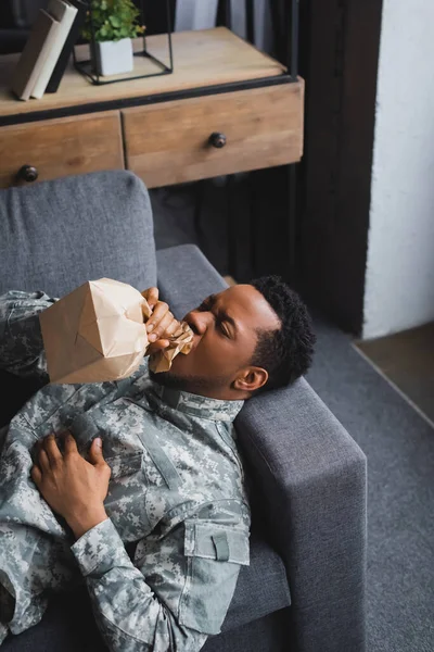 African american soldier in military uniform breathing with paper bag while having panic attack at home — Stock Photo
