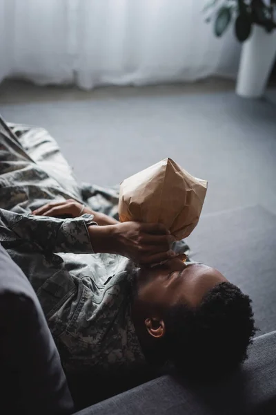 African american soldier in military uniform breathing with paper bag while having panic attack at home — Stock Photo