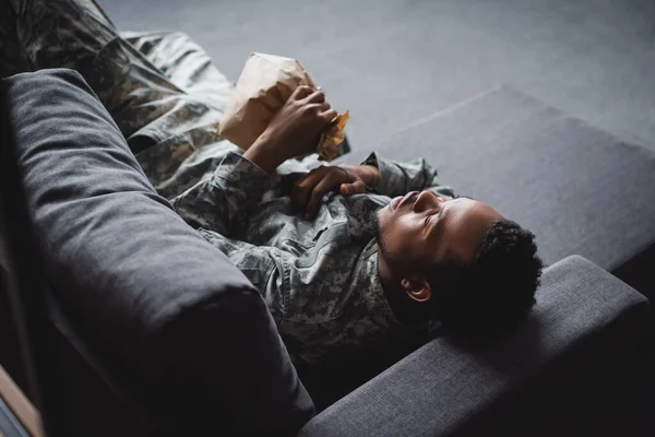 Soldado afroamericano en uniforme militar sosteniendo bolsa de papel mientras tiene ataque de pánico y sufrimiento de PTSD en casa - foto de stock