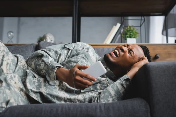 Stressed african american soldier crying while holding army badge and smartphone, suffering from PTSD at home — Stock Photo