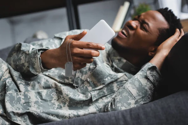 Stressed african american soldier crying while holding army badge and smartphone, suffering from PTSD at home — Stock Photo