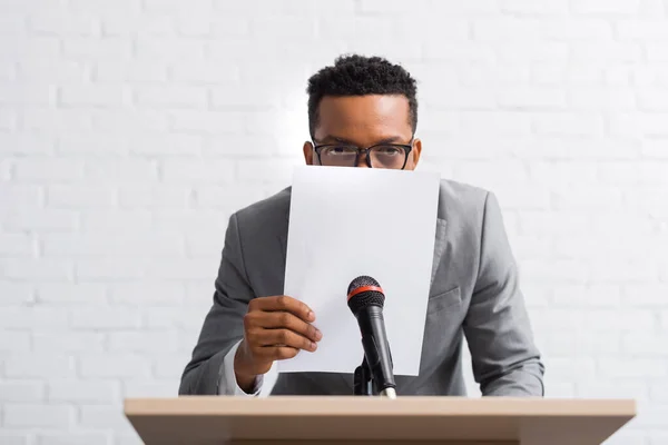 Nervous african american speaker hiding behind paper during business conference — Stock Photo