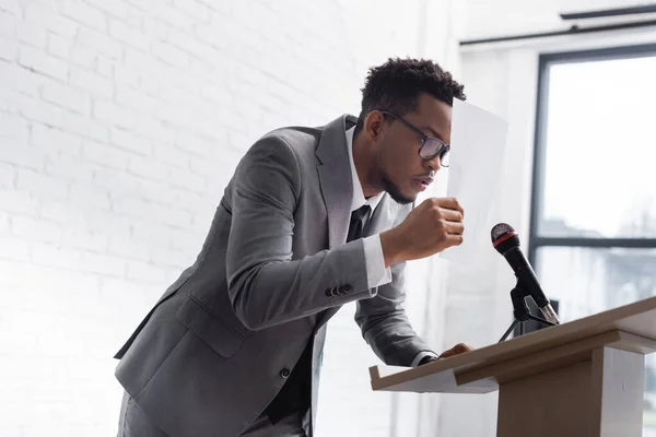 Stressed african american speaker hiding behind paper during business conference — Stock Photo