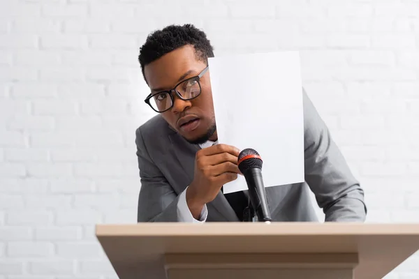 Scared african american speaker hiding behind paper during business conference — Stock Photo