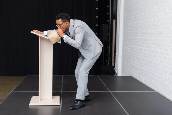 Acentuado empresario afroamericano respirando con bolsa de papel y teniendo ataque de pánico durante conferencia de negocios en la oficina - foto de stock