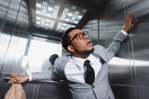 Scared african american businessman with paper bag suffering from panic attack in elevator — Stock Photo