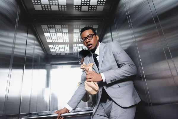 Nervioso hombre de negocios afroamericano con bolsa de papel que sufre de ataque de pánico en ascensor - foto de stock