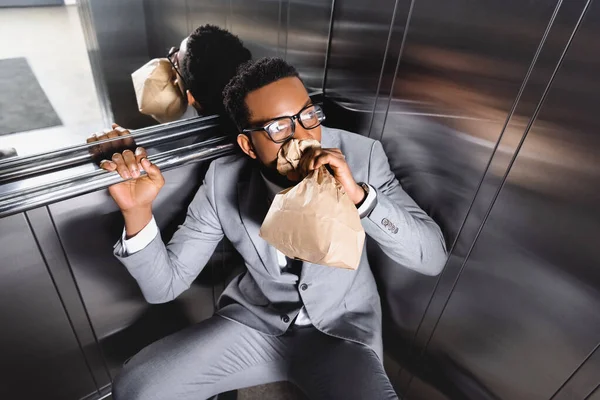 Hombre de negocios afroamericano asustado respirando con bolsa de papel y sufriendo de ataque de pánico en ascensor - foto de stock