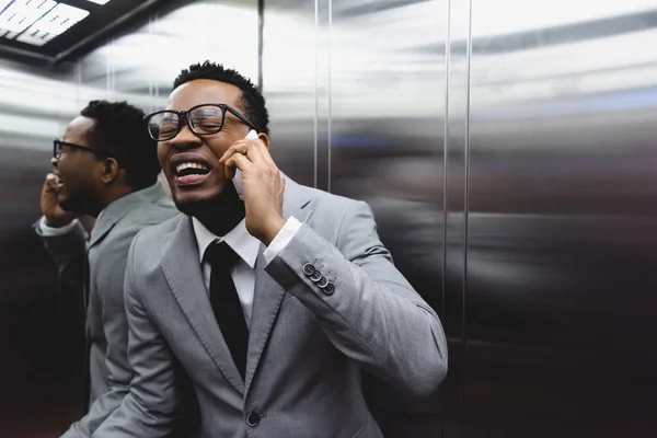 Crying african american businessman talking on smartphone and suffering from panic attack in elevator — Stock Photo