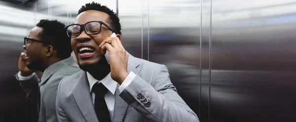 Panoramic shot of frightened crying african american businessman talking on smartphone and suffering from panic attack in elevator — Stock Photo