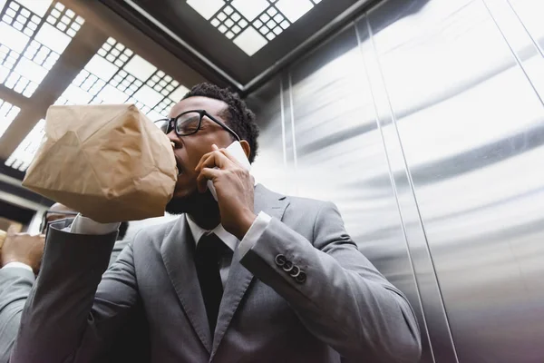 Stressed african american businessman talking on smartphone while breathing with paper bag and suffering from panic attack in elevator — Stock Photo