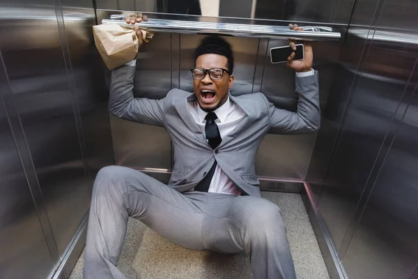 Scared screaming african american businessman with smartphone and paper bag suffering from panic attack in elevator — Stock Photo