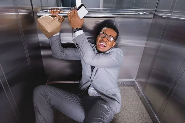 Homem de negócios afro-americano estressado com smartphone e saco de papel sofrendo de ataque de pânico no elevador — Fotografia de Stock